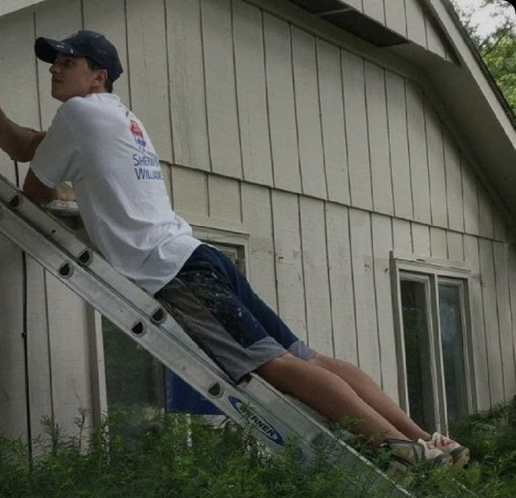 Early Wildwood painter leaning from a ladder to brush trim on a white sided house