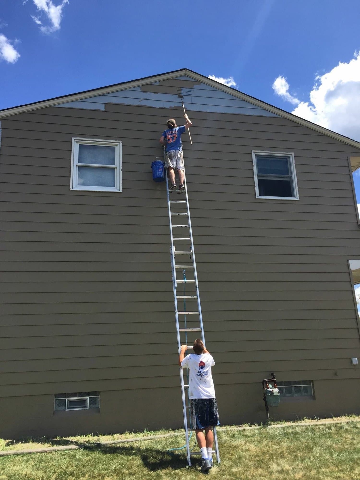 Two painters on ladders working on a two-story home, one rolling the gable from the top of the ladder