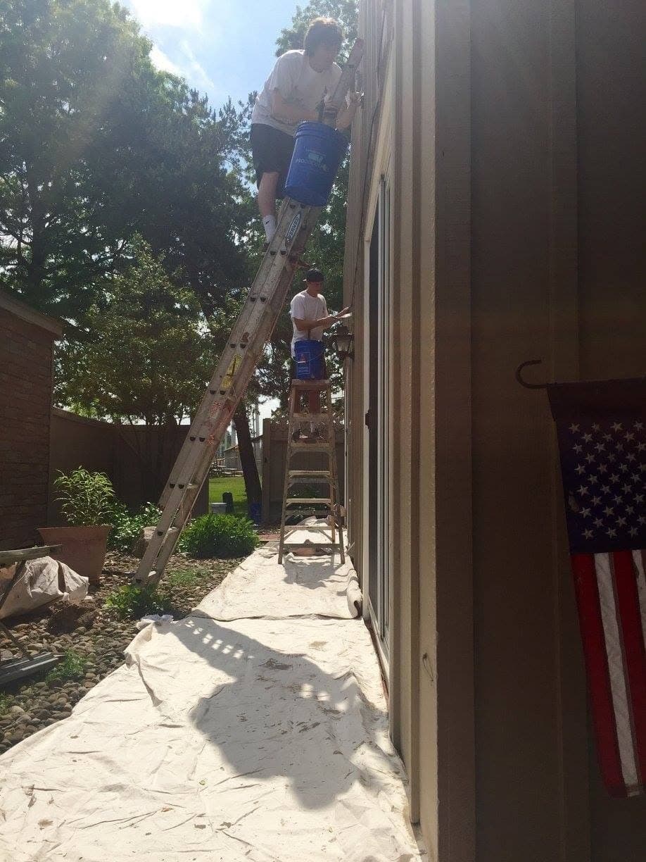 Two painters on a ladder alongside tall vertical siding, drop cloths on the path below