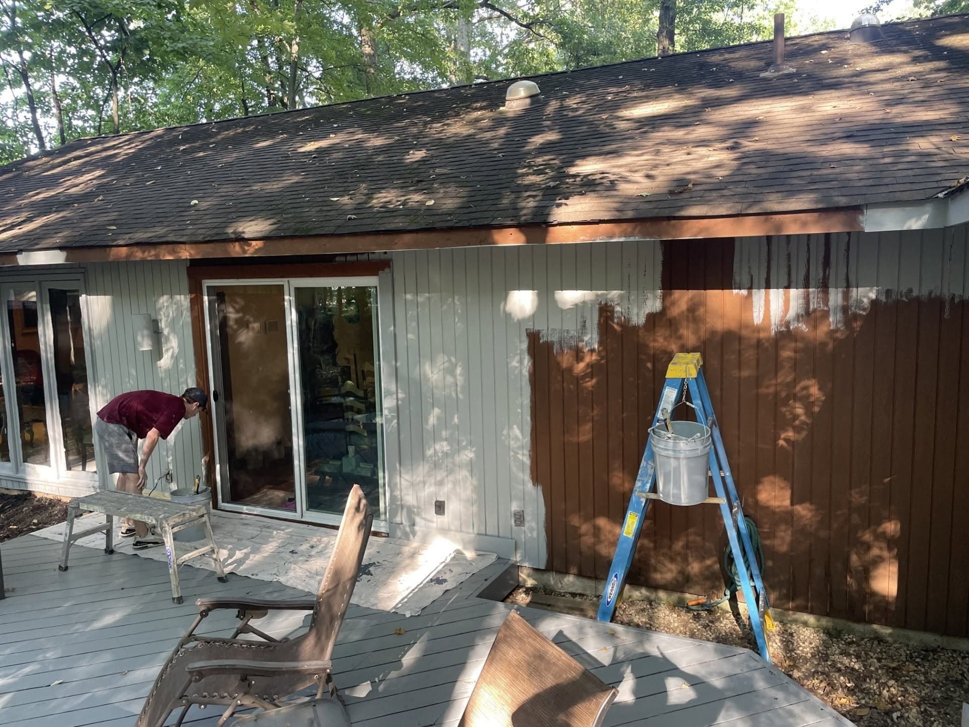Brown wood siding partway through being repainted white, with a ladder and paint bucket beside a back deck