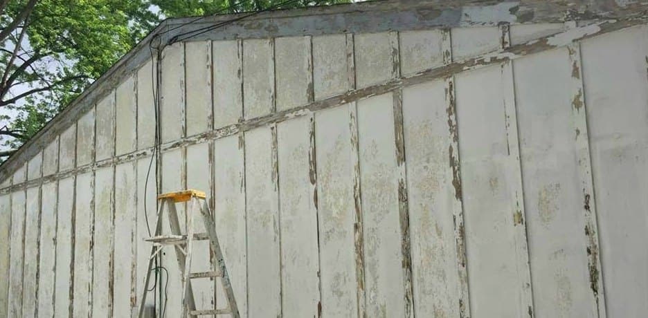 Board-and-batten siding with heavily peeling white paint and a ladder against the wall, before prep