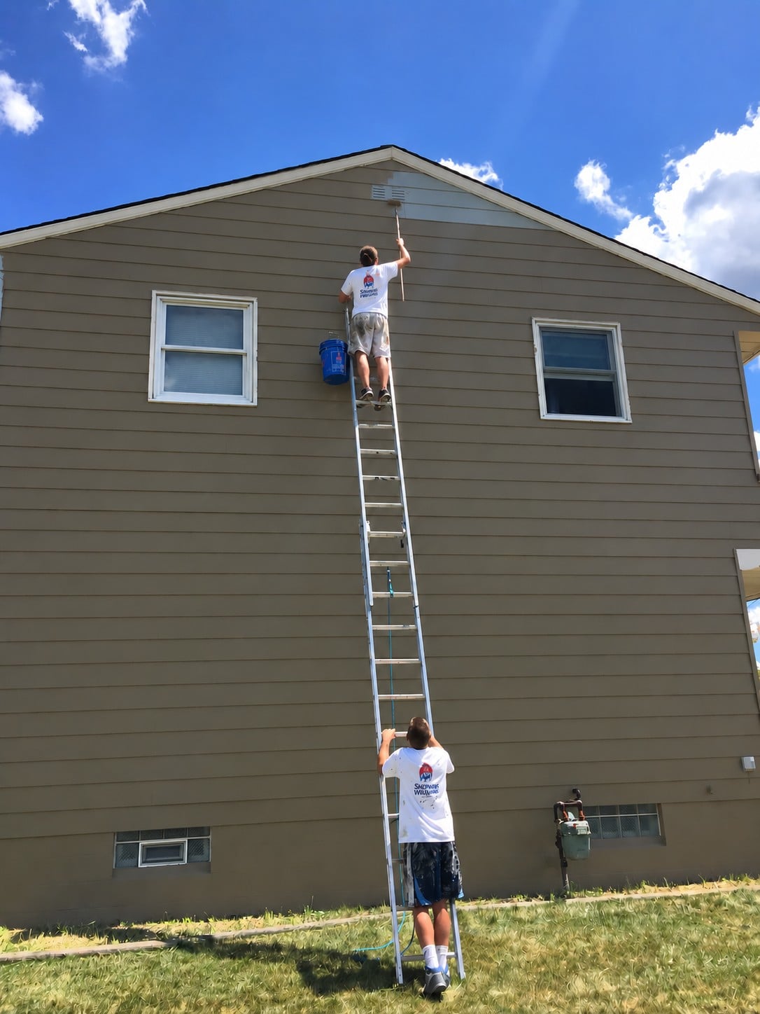 Two Wildwood painters on a ladder against a two-story home, one rolling paint onto the gable while the other steadies the ladder